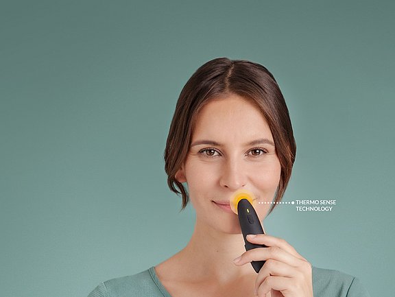 Eine Frau hält ein kleines schwarz-gelbes Gerät an ihre Oberlippe, auf dem vor einem schlichten blaugrünen Hintergrund der Text „THERMO SENSE TECHNOLOGY“ steht.